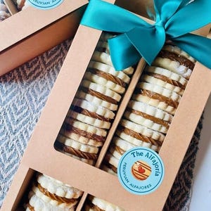 May include: A rectangular cardboard gift box with a clear window containing two rows of stacked round sandwich cookies filled with caramel. The box is tied with a teal ribbon and features a circular sticker labeled The Alfajoria Peruvian Alfajores.