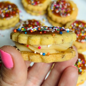 May include: A sandwich cookie with scalloped edges, featuring a light brown biscuit base, a smooth beige cream filling, and a glossy caramel-colored top layer decorated with small, multicolored round sprinkles, with more cookies visible in the background.