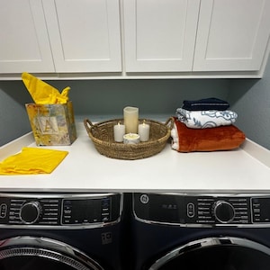 May include: A laundry room scene featuring a white countertop over two dark blue washing machines. On the counter are a tissue box, a woven basket with candles, and folded blankets. A yellow cloth is also present.