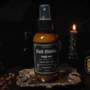 Puede incluir: Una botella de vidrio marr&oacute;n con una boquilla de pulverizaci&oacute;n negra y una etiqueta que dice "Dark Divinity magick mist". La botella est&aacute; sentada en una bandeja dorada con una vela y un libro en el fondo.