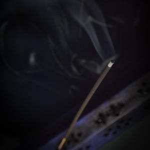 May include: A burning incense stick with smoke trails against a dark backdrop. The incense is brown with a white tip, and the smoke is wispy and white. The image has a soft focus, creating a calming atmosphere.