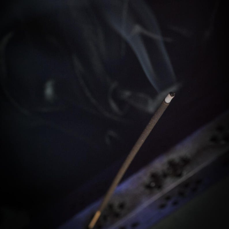 May include: A burning incense stick with smoke trails against a dark backdrop. The incense is brown with a white tip, and the smoke is wispy and white. The image has a soft focus, creating a calming atmosphere.