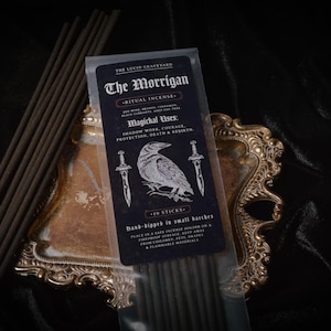 May include: A package of "The Morrigan" ritual incense with a black label featuring a raven illustration and text. The incense sticks are brown and are displayed on a decorative gold-colored tray.