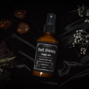 Puede incluir: Una botella de vidrio marr&oacute;n oscuro con una etiqueta negra que dice "Dark Divinity magic mist". La botella tiene una boquilla de pulverizaci&oacute;n plateada y est&aacute; sobre una superficie de tela negra.