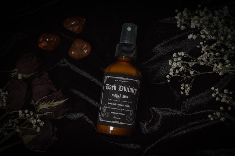 Puede incluir: Una botella de vidrio marr&oacute;n oscuro con una etiqueta negra que dice "Dark Divinity magic mist". La botella tiene una boquilla de pulverizaci&oacute;n plateada y est&aacute; sobre una superficie de tela negra.