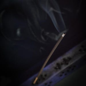 May include: A lit incense stick with smoke rising against a dark background. The incense is brown with a white tip, and the smoke is wispy and white. A decorative incense holder is visible in the lower right corner.