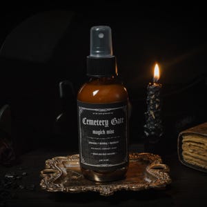 May include: A brown glass bottle of "Cemetery Gate magick mist" with a black label, set on a decorative gold tray. The label lists ingredients and the bottle has a black spray nozzle. A lit candle and a book are in the background.