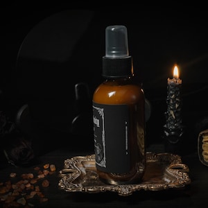 Puede incluir: Una botella de vidrio marr&oacute;n con una etiqueta negra y una boquilla de pulverizaci&oacute;n transparente. La botella est&aacute; sentada en una bandeja de metal dorado. Una vela negra est&aacute; ardiendo en el fondo.