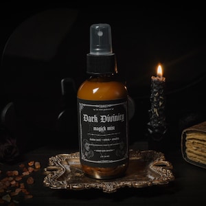 May include: A brown glass bottle labelled "Dark Divinity magick mist" with a black cap and spray nozzle. The bottle is on a decorative silver tray, with a lit candle and a book in the background. The label includes the text "shadow work + rebirth + rituals".