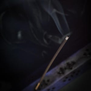 May include: A lit incense stick with smoke rising against a dark background. The incense is brown with a white tip, and the smoke is wispy. A decorative incense holder is visible in the lower right corner.
