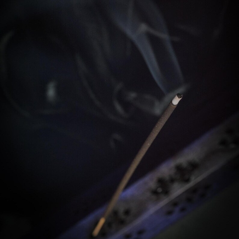May include: A lit incense stick with smoke rising against a dark background. The incense is brown with a white tip, and the smoke is wispy. A decorative incense holder is visible in the lower right corner.