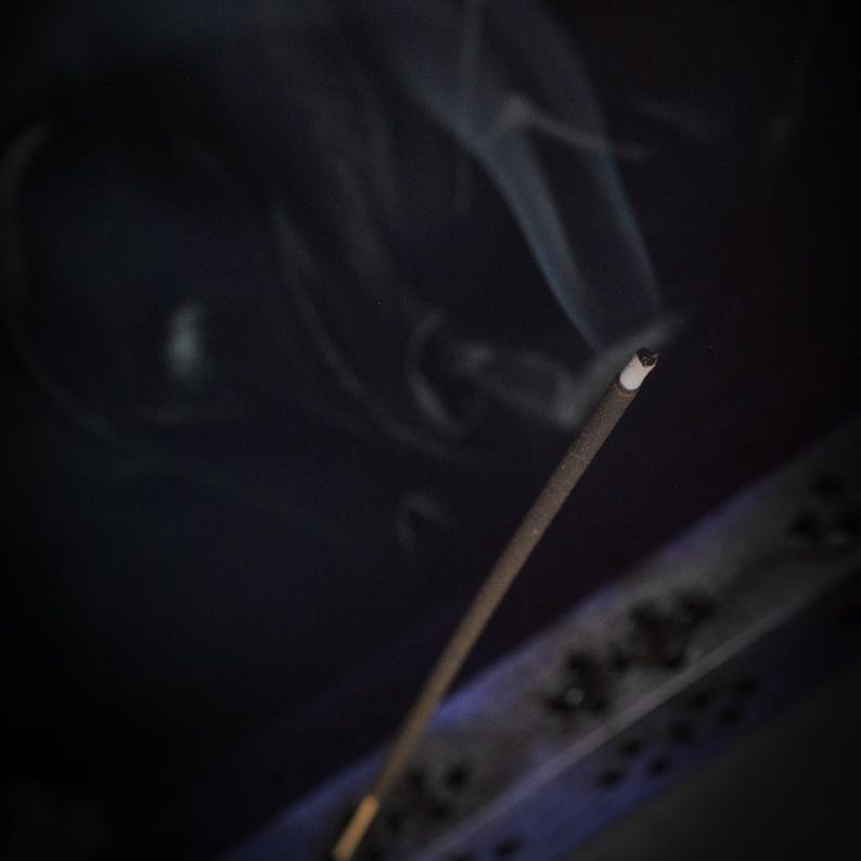 May include: A lit incense stick with smoke rising against a dark background. The incense is brown with a white tip, and the smoke is wispy and white. A decorative holder is visible in the lower right corner.