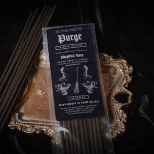 May include: A package of incense sticks labeled "Purge - Ritual Incense" with text detailing ingredients and uses. The package is on a gold-colored decorative tray, with additional incense sticks visible. The text includes "Remove Negative Energy, Cleansing, Purification, Banishment."