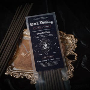 May include: Dark Divinity ritual incense sticks in a clear plastic package, with text that reads "The Lucid Graveyard". The package is on a gold-colored decorative tray, with several incense sticks visible. The text describes the incense's magical uses.