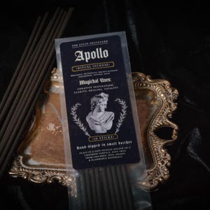 May include: A package of "Apollo" ritual incense sticks, hand-dipped in small batches. The package is dark blue with white text and a bust illustration. The incense sticks are dark brown and rest on a decorative gold-colored tray.