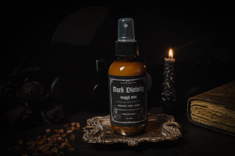Puede incluir: Una botella de vidrio marr&oacute;n con una boquilla de pulverizaci&oacute;n negra y una etiqueta que dice "Dark Divinity magick mist". La botella est&aacute; sentada en una bandeja dorada con una vela y un libro en el fondo.