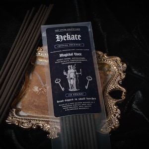 May include: A package of Hekate ritual incense with a dark blue label, featuring text and illustrations. The package is on a decorative gold-colored tray, with several incense sticks visible. The text includes "Magichal Uses: Spirit Work, Divination, Witchcraft, Empowerment, Transformation."