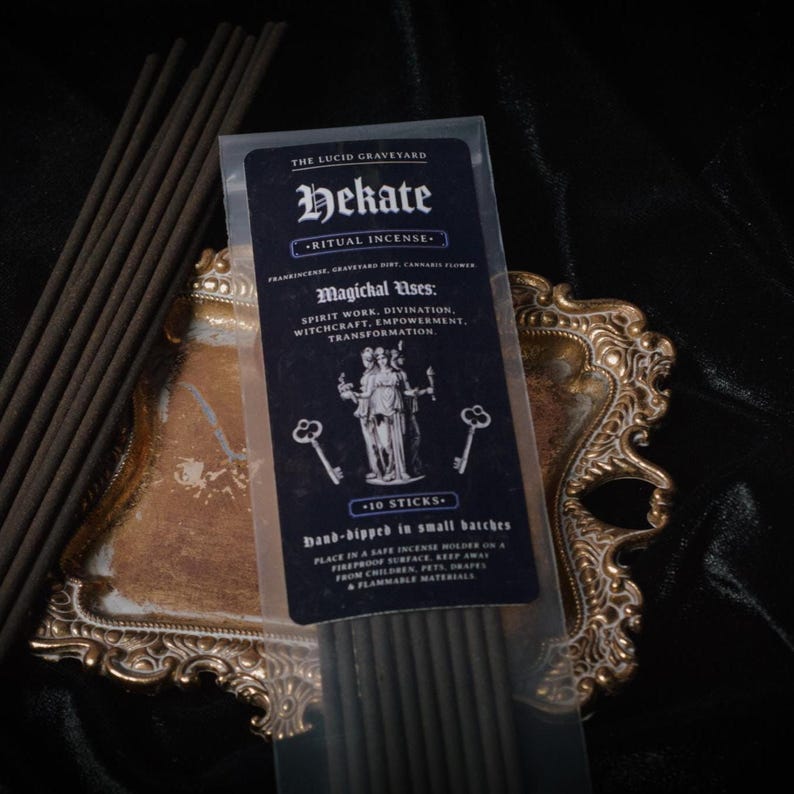 May include: A package of Hekate ritual incense with a dark blue label, featuring text and illustrations. The package is on a decorative gold-colored tray, with several incense sticks visible. The text includes "Magichal Uses: Spirit Work, Divination, Witchcraft, Empowerment, Transformation."