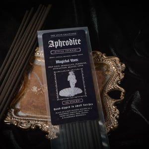 May include: A package of Aphrodite ritual incense sticks with a black and white label, featuring an illustration of a woman. The incense is displayed on a decorative gold-colored tray against a dark background.
