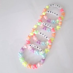 May include: Five colourful beaded bracelets with names spelled out in white letter beads: DAISY, BELLA, JESS, ROSE, and WINNIE. The bracelets feature pastel-coloured beads and flower-shaped accents, with a white background.