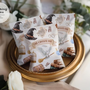 May include: White treat bags with a watercolor design featuring a black witch hat, owl, books, and the text "THE CHOSEN ONE." The bags are displayed on a gold-rimmed tray, with a Canva logo in the corner.