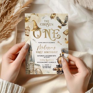 May include: A gold and white invitation card with a watercolor illustration of a train, a castle, and a wizard's hat. The text on the card reads "The Chosen One" and "Melissa's First Birthday!"