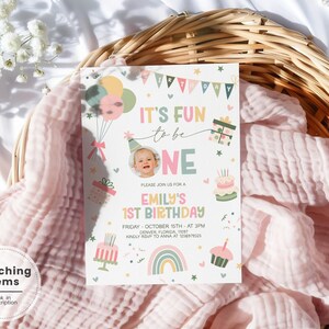 May include: A rectangular birthday invitation card featuring a photo of a baby, pastel-colored balloons, a rainbow, cakes, and gift boxes. The text reads: It's fun to be one, Emily's 1st Birthday, Friday October 15th at 3pm, Denver Florida 11091, kindly RSVP to Anna at 1234567890.