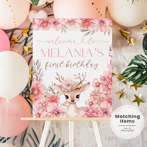 Peut inclure: Panneau de bienvenue pour le premier anniversaire de Melania, avec une illustration de cerf et des fleurs roses. Le texte dit "welcome to MELANIA'S first birthday". Des ballons roses, blancs et dorés complètent la décoration.