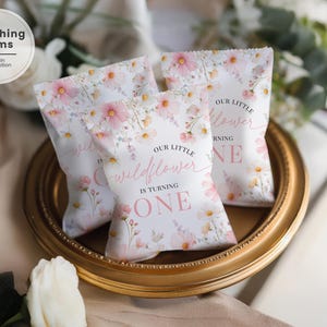 May include: Three white favor bags with a floral design of pink and yellow flowers, displayed on a gold-rimmed tray. The bags have text that reads "Our little wildflower is turning one."