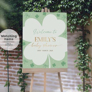 Może przedstawiać: Znak baby shower na drewnianym sztaludze. Znak jest jasnozielony z dużym białym kształtem koniczyny. Złoty tekst brzmi "Witamy na baby shower EMILY 15 marca 2025". Ozdobne koniczyny i kokardy również znajdują się na znaku.