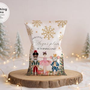 May include: A white gift bag with gold snowflakes and the text "Our little Sugar plum is turning one!" features illustrations of a nutcracker, ballerina, and mouse king. The bag is displayed on a wooden slice with fairy lights.