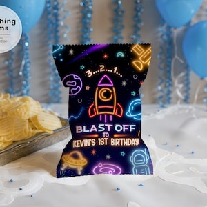 May include: A black chip bag with a neon space-themed design. The bag features a rocket, planets, and the text "BLAST OFF TO KEVIN'S 1ST BIRTHDAY." Blue balloons and chips are in the background.