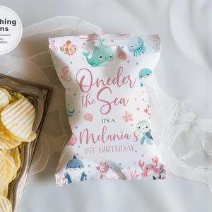 May include: A white chip bag with an "Oneder the Sea" theme, featuring sea creatures and the text "It's a Melania's 1st Birthday." The bag is decorated with cartoon whales, fish, and other marine life. A silver tray of chips is in the foreground.