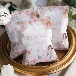 May include: Three white party favor bags with a floral and fairy design, arranged on a gold-colored tray. The bags are decorated with pink flowers, butterflies, and illustrations of fairies. Text on the bags reads "MELISSA'S fairy first birthday."