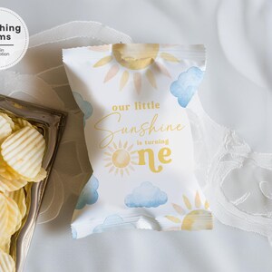 May include: A white snack bag featuring a yellow sun illustration and blue watercolor clouds with the text our little Sunshine is turning one printed in yellow and gold script and sans-serif fonts, positioned next to a silver tray of potato chips.
