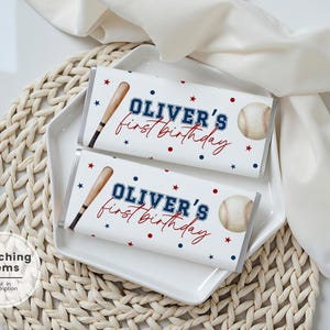 May include: Two white candy bar wrappers with baseball designs. Each wrapper has a baseball bat, baseball, and the text "OLIVER'S first birthday" in blue and red. The wrappers are on a white plate.