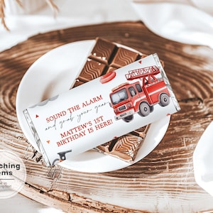 Puede incluir: Envoltorio de barra de chocolate blanco con una ilustración de camión de bomberos rojo y el texto "SOUND THE ALARM and grab your gear. MATTEW'S 1ST BIRTHDAY IS HERE!". La barra está sobre un plato blanco, sobre una superficie de madera.