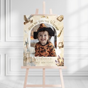 K&ouml;nnte beinhalten: Eine h&ouml;lzerne Staffelei zeigt ein individuelles Geburtstagszeichen mit einem Kinderfoto. Das Schild hat ein Zaubererthema mit dem Text "Have You Seen This Wizard?" und "Melissa's First Birthday!". Dekorative Elemente sind ein Schloss, eine Eule und Kristalle.