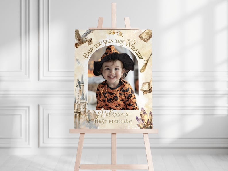 K&ouml;nnte beinhalten: Eine h&ouml;lzerne Staffelei zeigt ein individuelles Geburtstagszeichen mit einem Kinderfoto. Das Schild hat ein Zaubererthema mit dem Text "Have You Seen This Wizard?" und "Melissa's First Birthday!". Dekorative Elemente sind ein Schloss, eine Eule und Kristalle.
