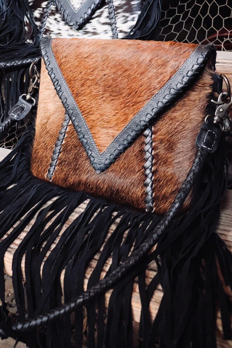Puede incluir: Un bolso de mano de cuero marr&oacute;n con flecos negros y una correa trenzada. El bolso tiene un dise&ntilde;o triangular &uacute;nico con costuras negras.