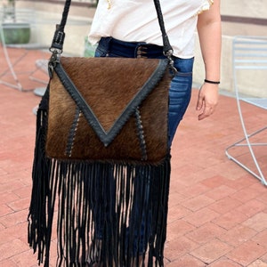 Puede incluir: Un bolso bandolera de piel de vaca marr&oacute;n y negro con una larga franja negra. El bolso tiene cierre de solapa y una correa de cuero negro.