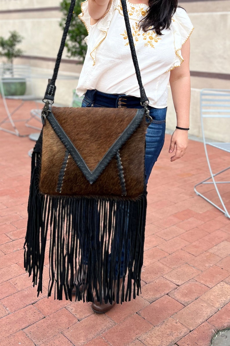 Puede incluir: Un bolso bandolera de piel de vaca marr&oacute;n y negro con una larga franja negra. El bolso tiene cierre de solapa y una correa de cuero negro.