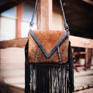 Puede incluir: Un bolso de cuero marr&oacute;n cruzado con flecos negros y una correa de cuero negro. El bolso tiene un dise&ntilde;o geom&eacute;trico en la parte delantera.