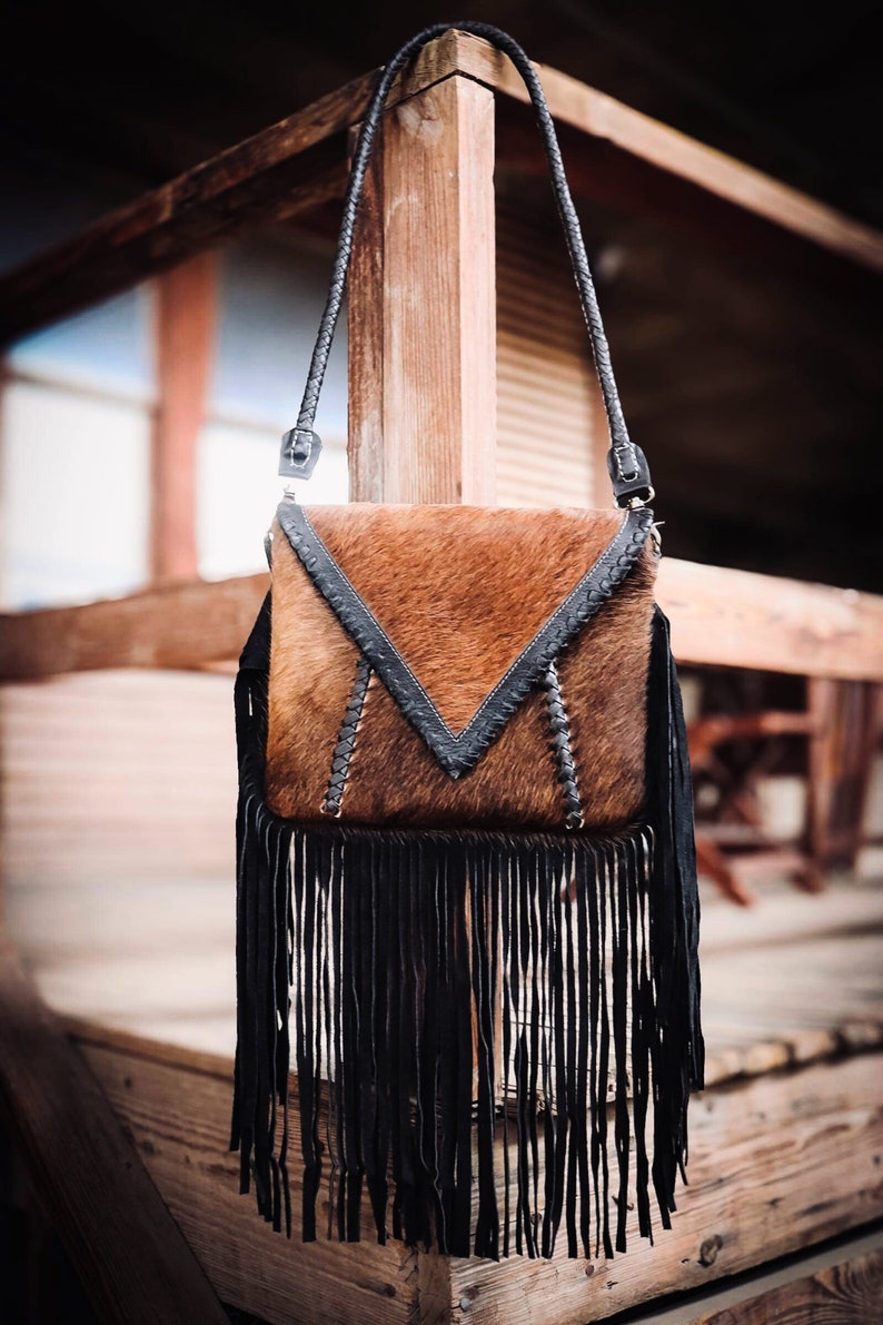 Puede incluir: Un bolso de cuero marr&oacute;n cruzado con flecos negros y una correa de cuero negro. El bolso tiene un dise&ntilde;o geom&eacute;trico en la parte delantera.
