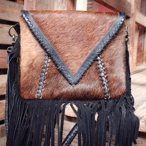 Puede incluir: Un bolso bandolera de piel de vaca marr&oacute;n y negro con flecos negros y una correa de cuero trenzada.