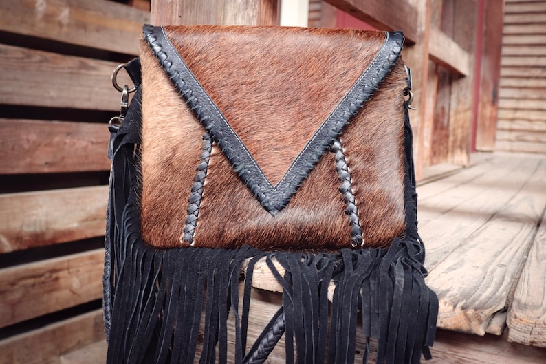 Puede incluir: Un bolso bandolera de piel de vaca marr&oacute;n y negro con flecos negros y una correa de cuero trenzada.