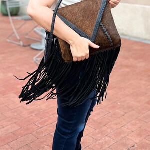 Puede incluir: Un bolso de mano de cuero marr&oacute;n y negro con detalles de flecos. El bolso tiene forma triangular y cierre de solapa.