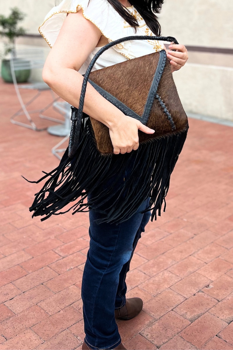 Puede incluir: Un bolso de mano de cuero marr&oacute;n y negro con detalles de flecos. El bolso tiene forma triangular y cierre de solapa.