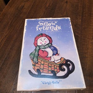 Pode incluir: Uma caixa retangular de produto para a série de moldes de biscoitos Snow Friends de 1998 da Longaberger Pottery. A caixa apresenta um fundo azul com uma ilustração branca de um boneco de neve com gorro vermelho e cachecol verde num trenó castanho, rotulado como Sleigh Belle.
