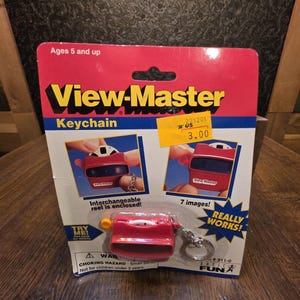 May include: A red View-Master keychain in its original cardboard packaging. The packaging features yellow and black text, including the words View-Master, Keychain, and Really Works. It displays two images of the product and a yellow price sticker.